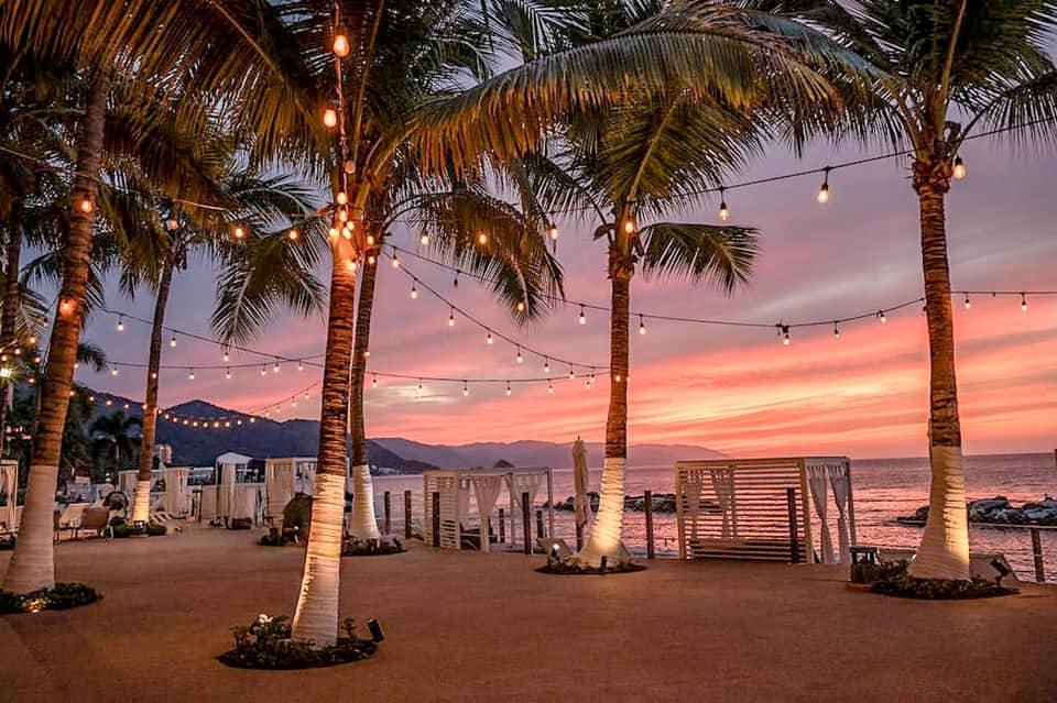 Beach venue at sunset with palm trees, string lights, wooden barriers, and colorful sky