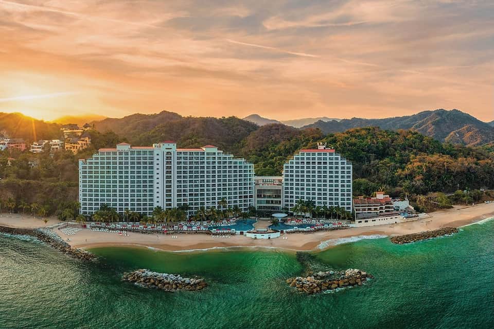 Beachfront high-rise hotel at sunset with ocean view, sandy beach, palm trees, and mountain backdrop