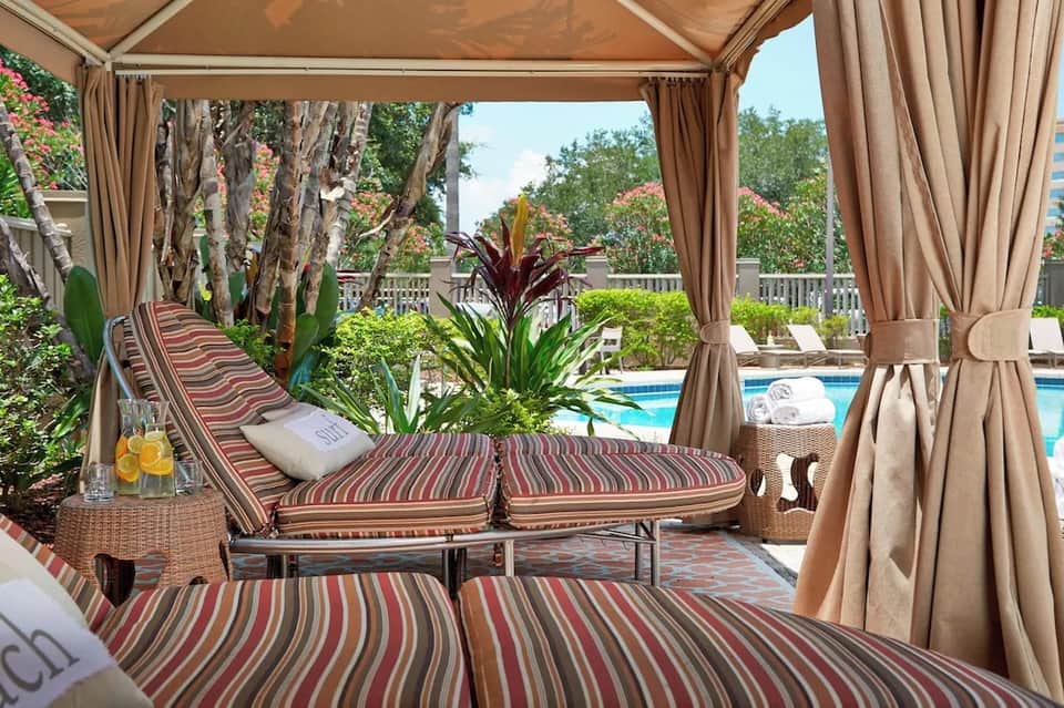 Luxurious poolside cabana with striped loungers, sheer curtains, tropical plants, and pool view