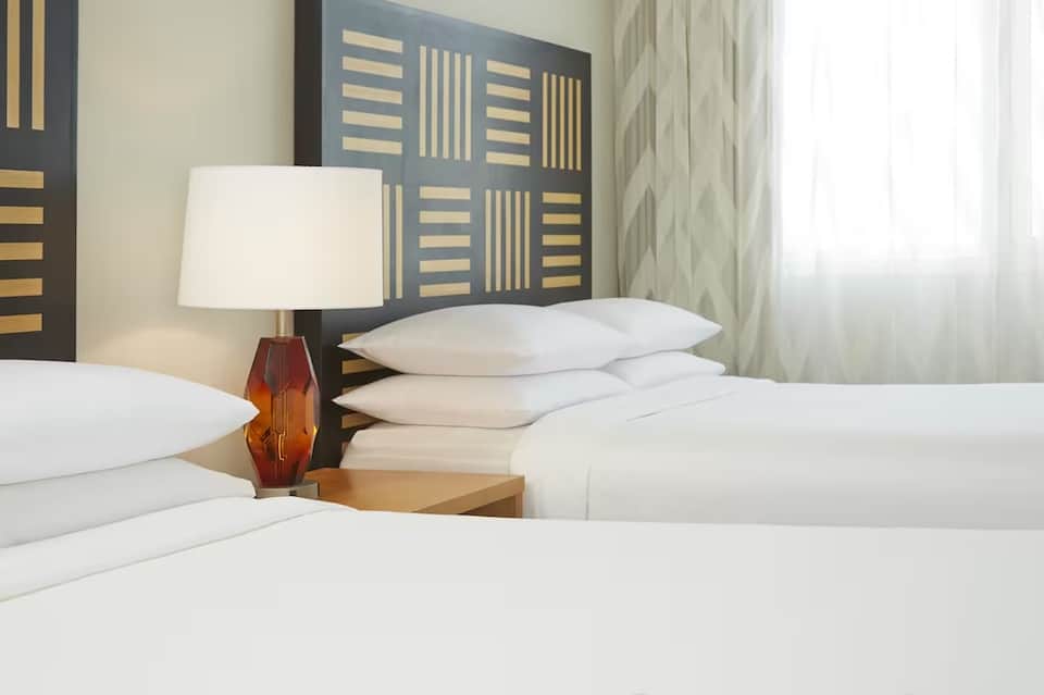 Hotel room detail showing white pillows, decorative headboard, bedside lamp, and curtains