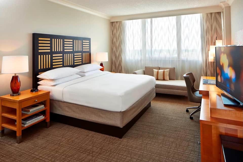 Modern hotel bedroom with queen bed, wooden headboard, desk, and floor-to-ceiling windows with city view