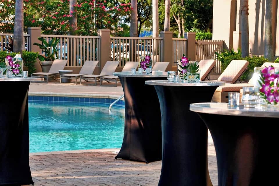 Upscale poolside event space with dining tables, lounge chairs, purple flowers, pool view, and palm trees