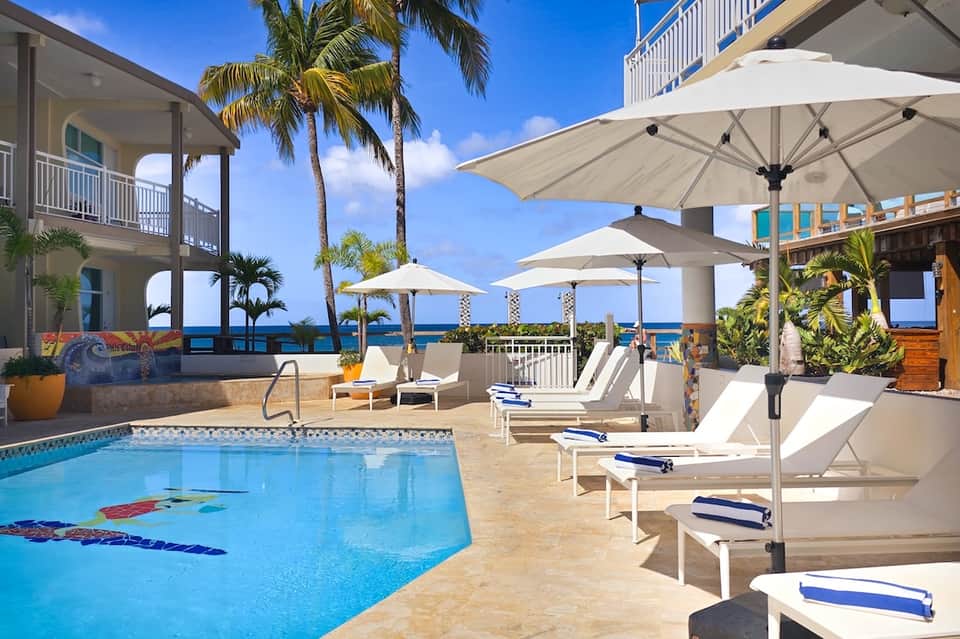 Oceanfront resort pool with white umbrellas, lounge chairs, palm trees, and ocean views