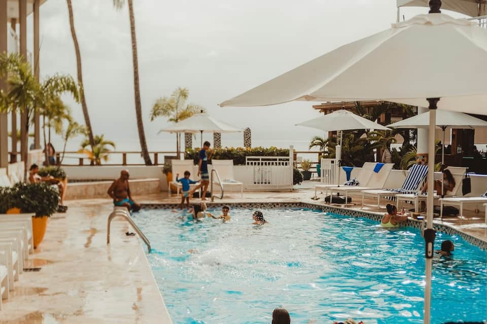 Beachfront pool with families swimming, white umbrellas, and palm trees at sunset