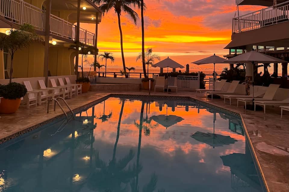 Oceanfront resort pool at golden sunset with palm trees, loungers, and beach umbrellas reflecting in water