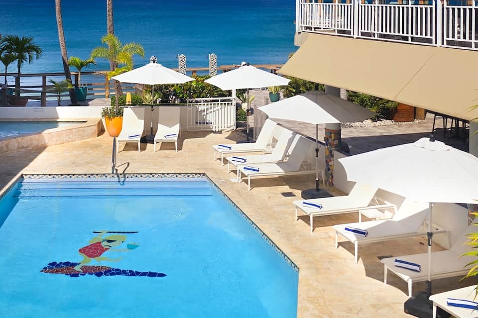 Beachfront pool with decorative fish mosaics, white lounge chairs, umbrellas, and ocean backdrop