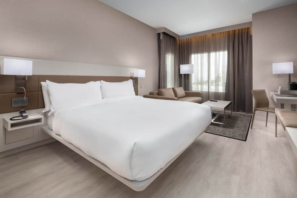 Modern hotel bedroom with king bed, seating area, and floor-to-ceiling windows