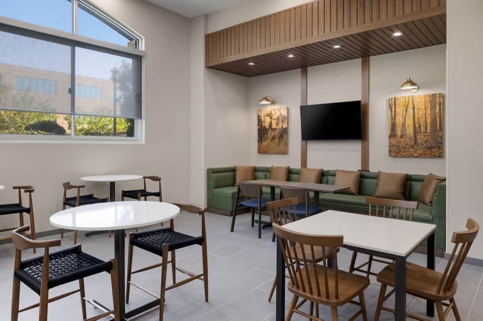 Modern hotel lounge with round tables, wooden chairs, green seating area, TV, and large windows
