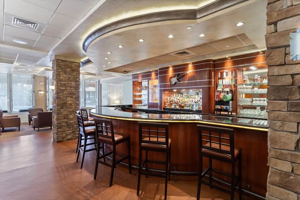 Hotel bar with curved dark wood counter, bar stools, stone pillar, ambient lighting, and view of lobby lounge area