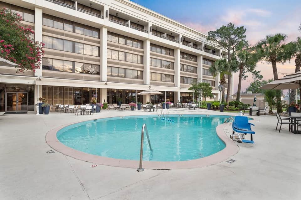 Modern resort pool with turquoise water surrounded by white concrete, lounge chairs, multi-story building facade, and palm trees