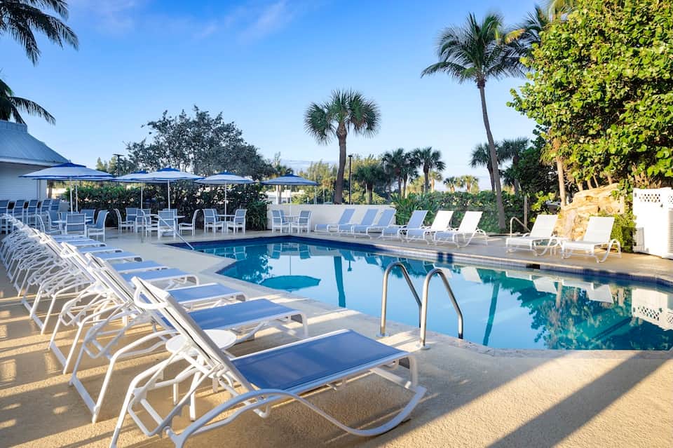 Resort pool area with white loungers, blue umbrellas, palm trees, and manicured tropical grounds