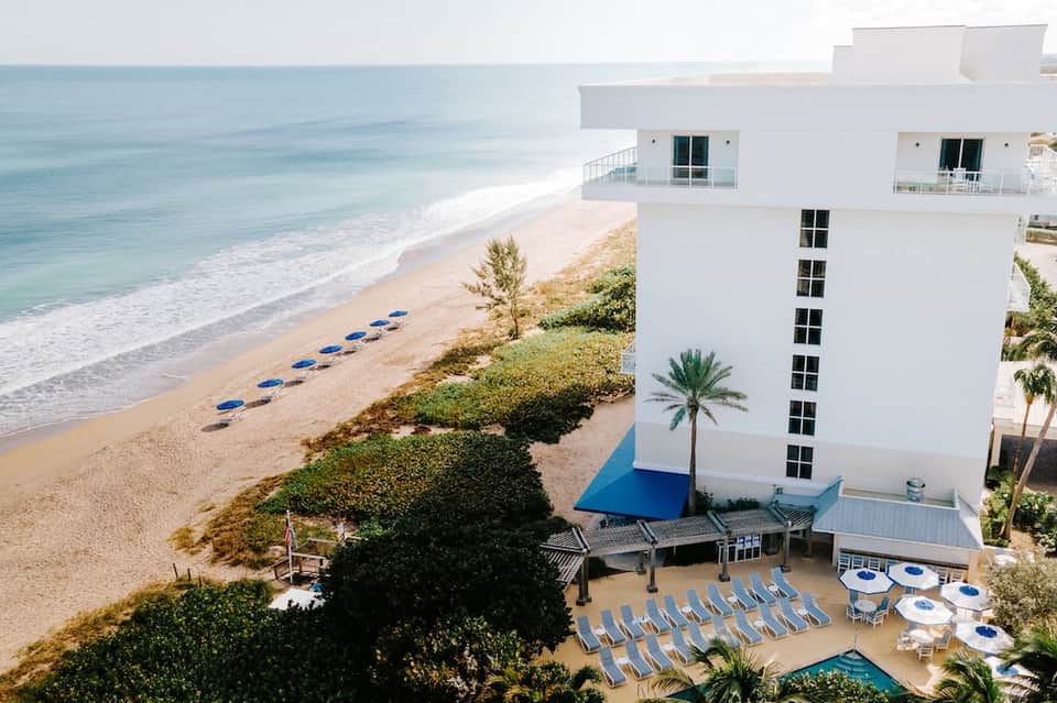 Beachfront resort building overlooking sandy beach, ocean, blue lounge chairs, white umbrellas, and palm landscaping