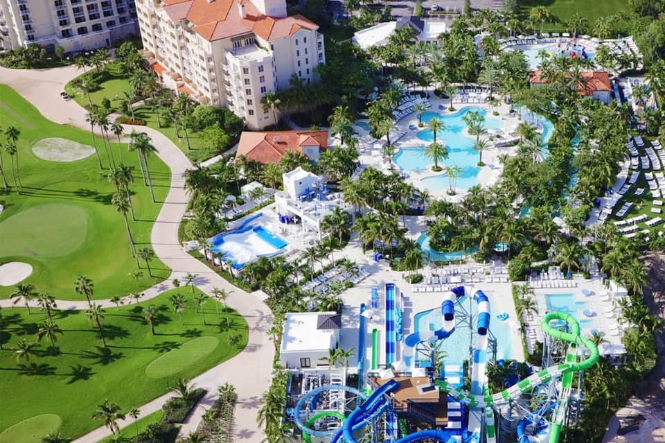 Aerial view of a resort with multiple swimming pools, water slides, palm trees, and surrounding buildings