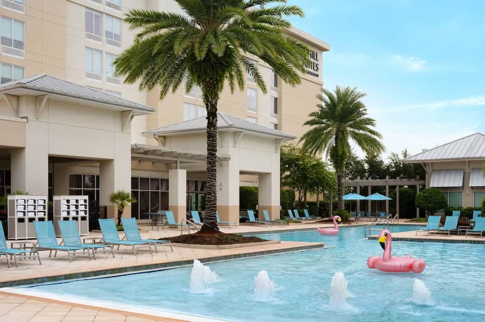Resort pool area with palm trees, lounge chairs, flamingo float, and hotel building