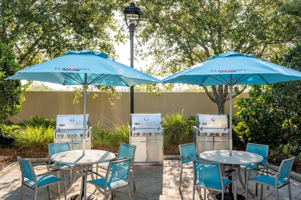 Patio area with grills under blue Flamingo umbrellas, turquoise chairs, and mature oak trees.