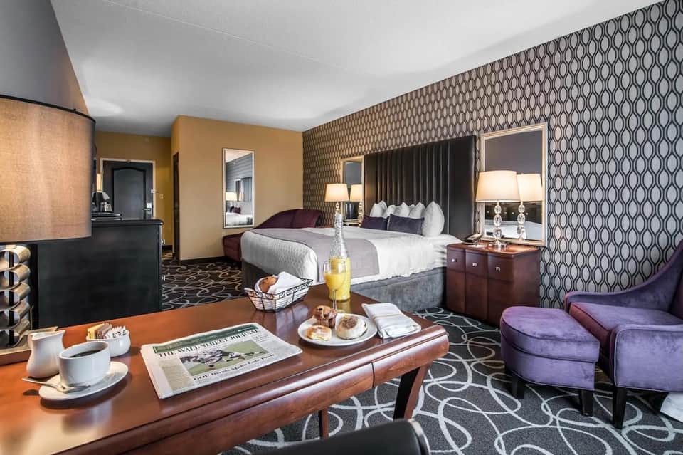 Luxury hotel suite with king bed, sitting area, patterned wallpaper, wooden desk, and purple accent chairs