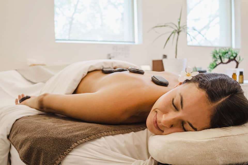 Woman receiving hot stone spa massage treatment on white bed in serene wellness room with windows.