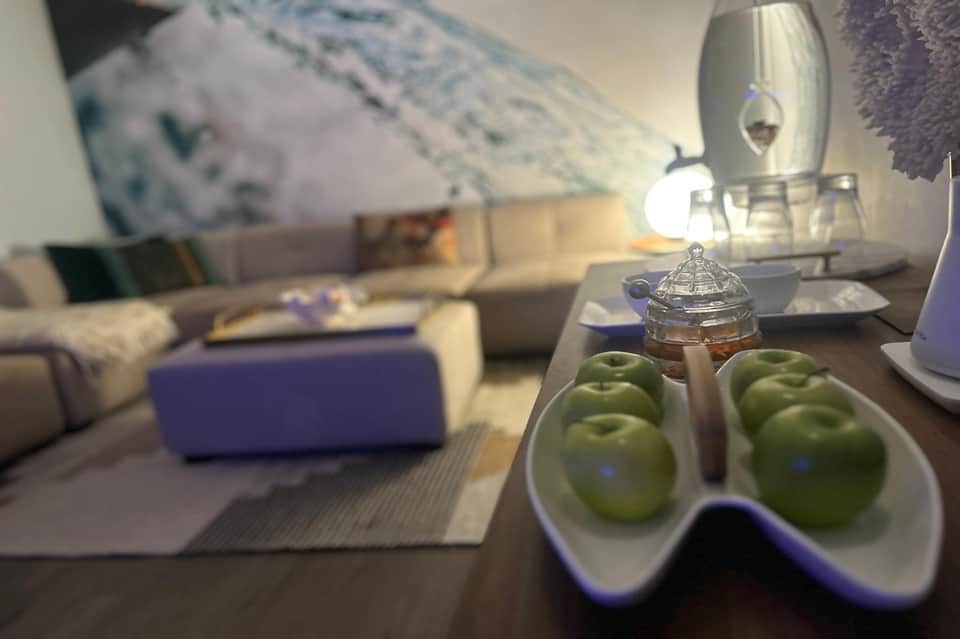 Hotel suite detail showing fresh green apples in bowl, glass container, and blurred seating area in background