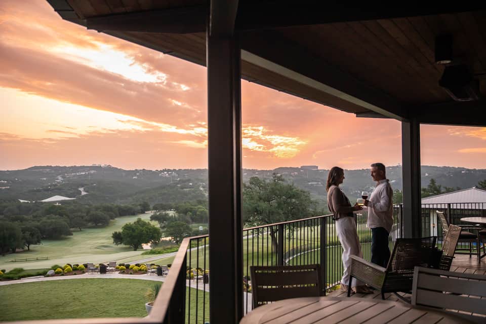 Couple on sunset balcony overlooking green valley, lake, and rolling hills at dusk