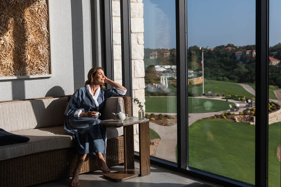 Woman in robe on seating area with expansive windows overlooking green hillside landscape and distant valley views