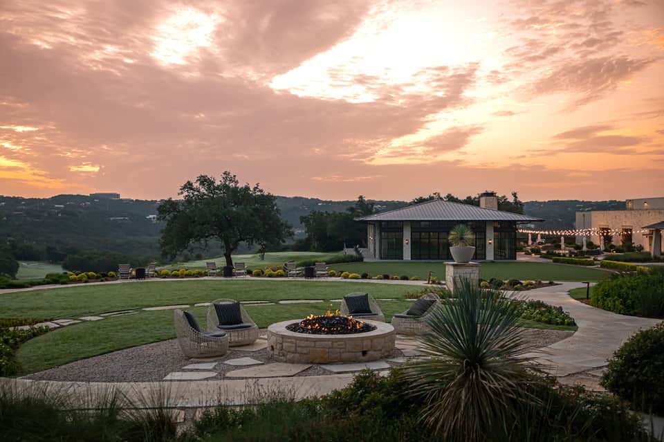 Sunset fire pit with woven chairs on stone patio overlooking manicured lawn and scenic valley landscape.