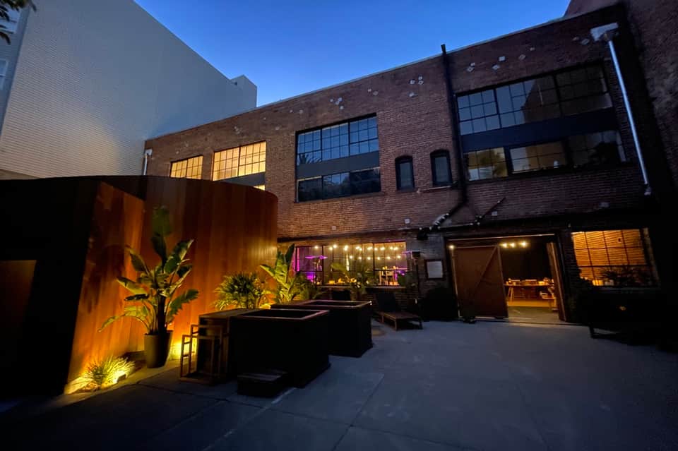 Urban hotel courtyard at dusk with ambient lighting, potted plants, and brick industrial-style building