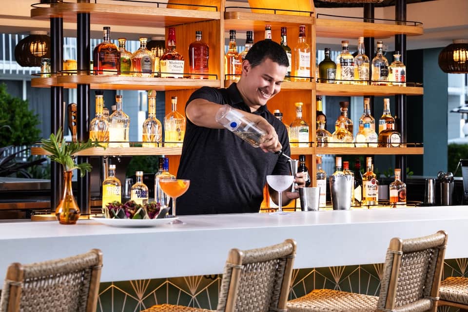Bartender crafting cocktail at modern bar with illuminated wooden shelving displaying liquor bottles