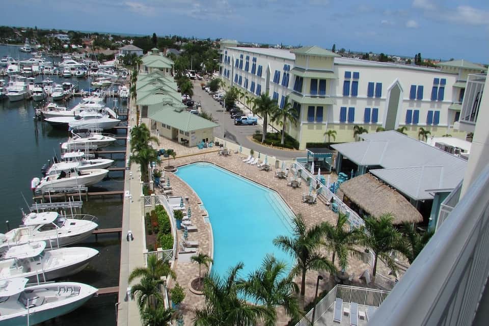 Waterfront resort aerial view with swimming pool, marina with boats, palm trees, and blue-roofed buildings