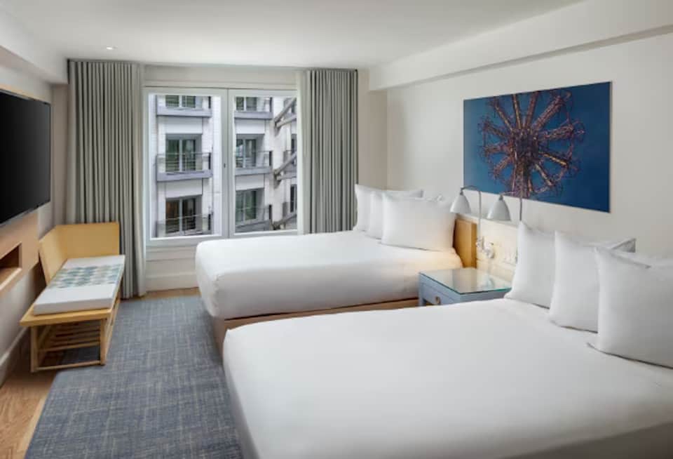 Hotel room with two queen beds, wooden headboards, modern decor, and city views through tall windows