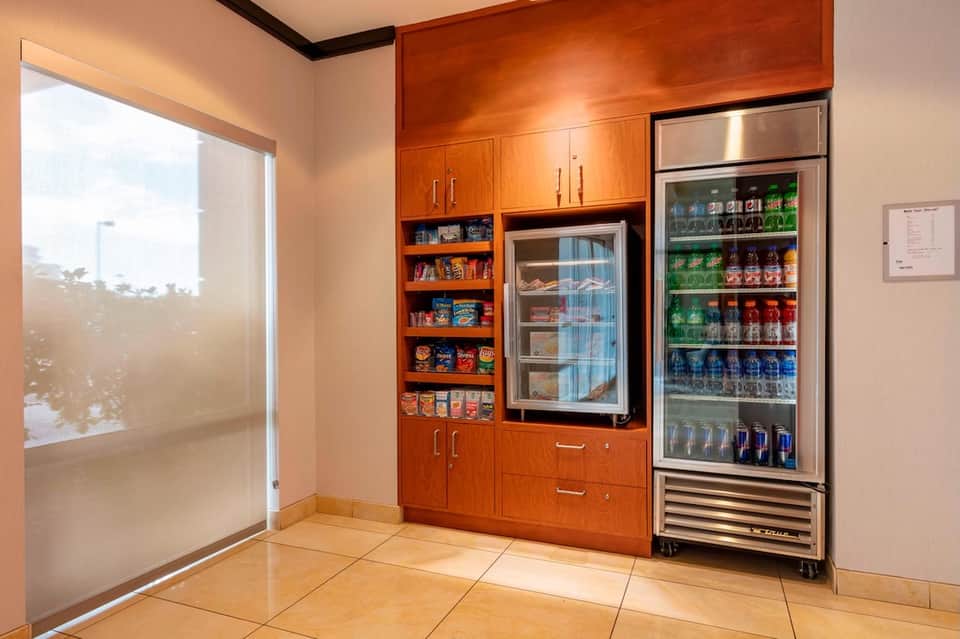 Hotel beverage station with wooden cabinetry, refrigerated displays, snacks, and frosted glass door