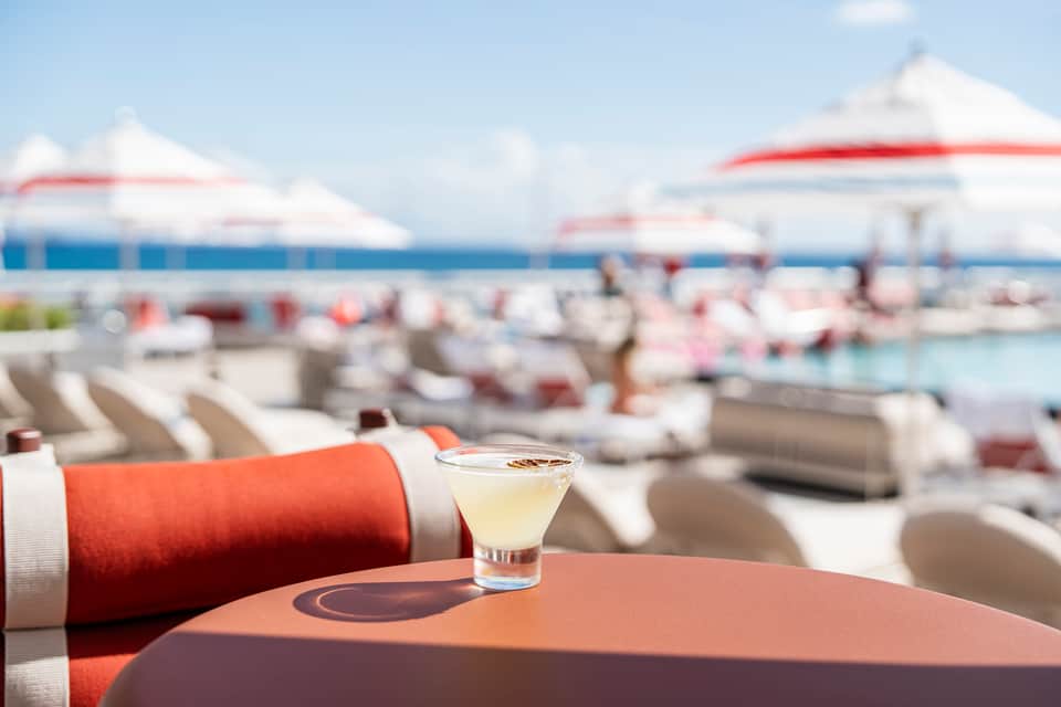 Cocktail glass on table with beach cabanas, umbrellas, and ocean view in background