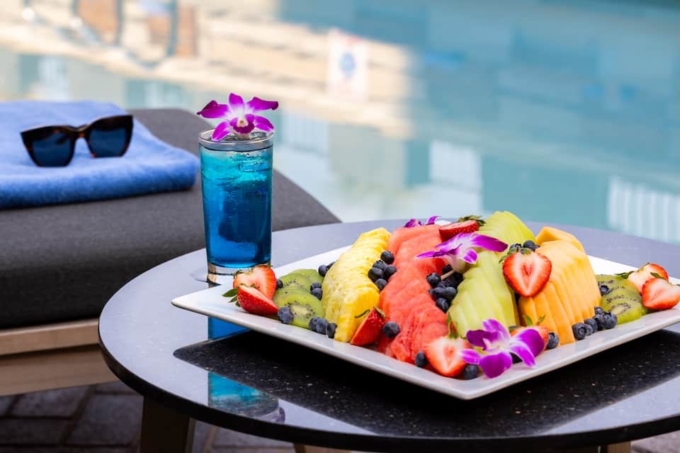 Poolside table with colorful fruit plate, blue cocktail, pink orchid, and city skyline backdrop
