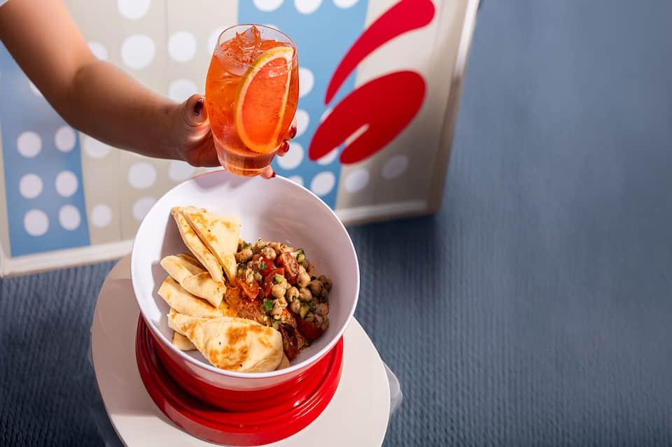 Hand holding orange cocktail above plate of hummus and pita chips at resort