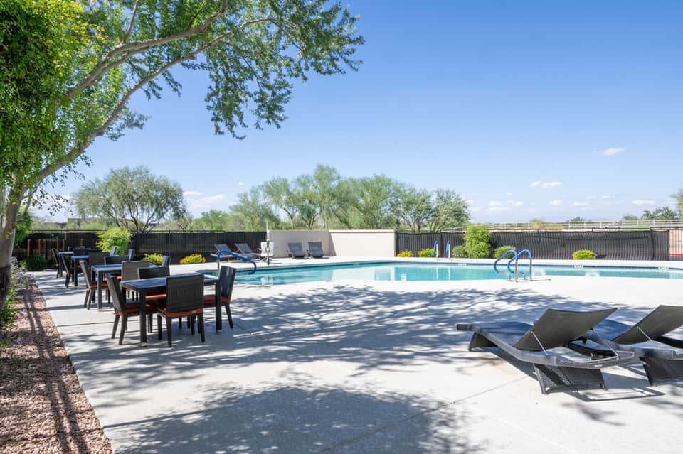 Resort pool area with shaded seating, lounge chairs, mature trees, and fenced grounds
