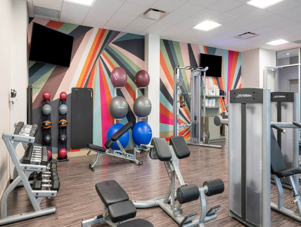Fitness center with colorful geometric mural, dumbbells, exercise equipment, and TVs