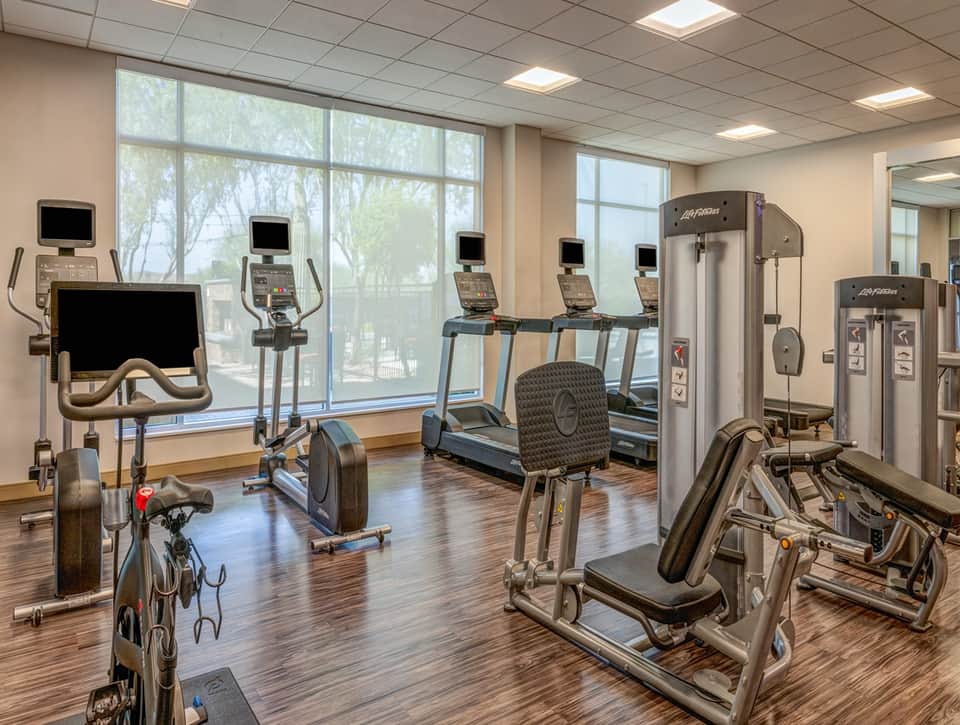 Bright fitness room with cardio equipment, exercise machines, windows overlooking trees, and wood flooring