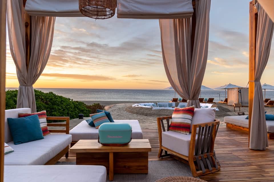 Luxury beachfront lounge with flowing curtains, sunset ocean view, wooden furniture, and colorful cushions on deck