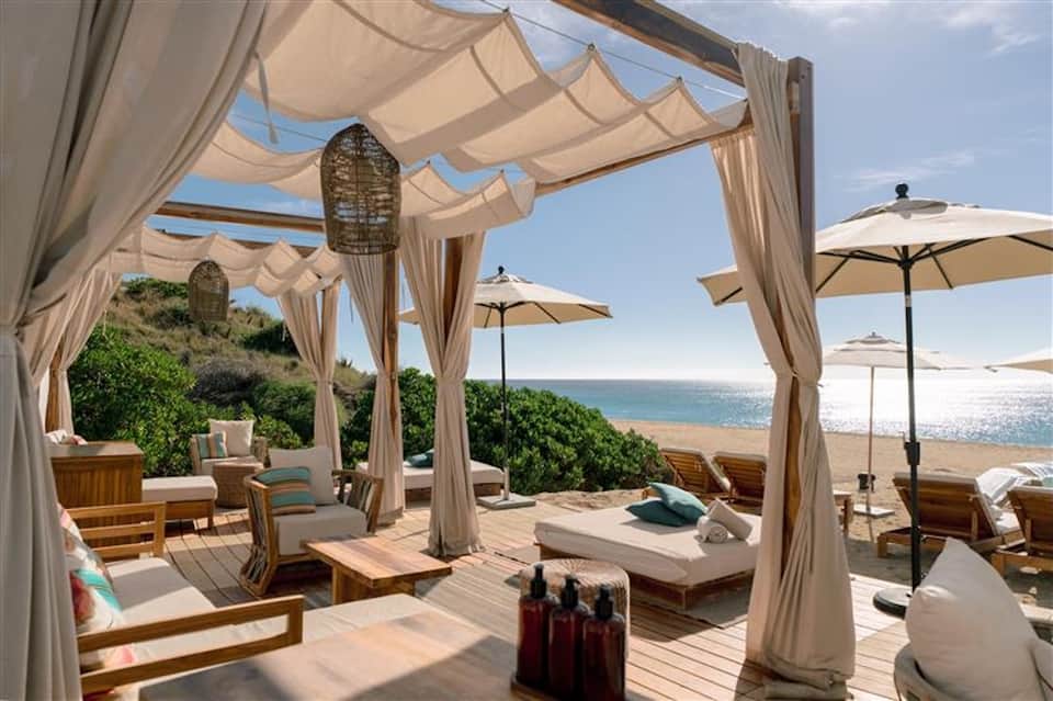 Beachfront resort terrace with wooden loungers, cream canopy, umbrellas, and ocean view