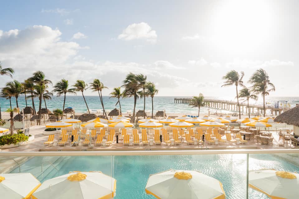 Beachfront resort pool terrace with yellow umbrellas, palm trees, ocean pier, and ocean views