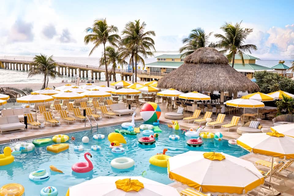 Beachfront resort pool with colorful floats, yellow umbrellas, palm trees, and tropical setting