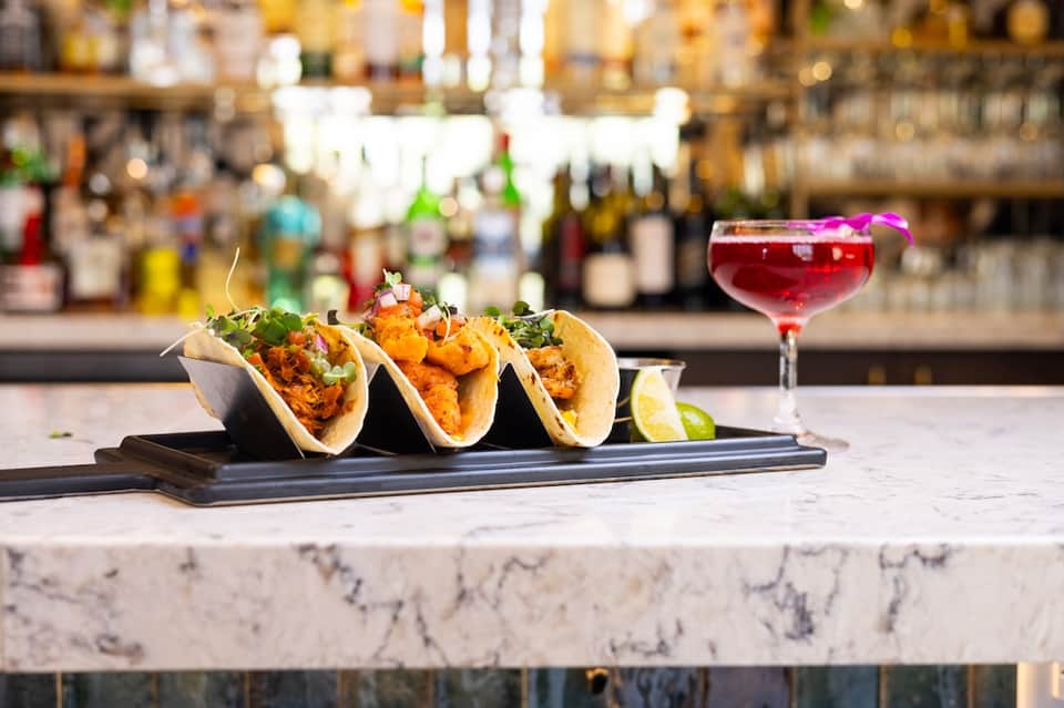 Three fish tacos on black plate with red cocktail at hotel bar with liquor bottles background