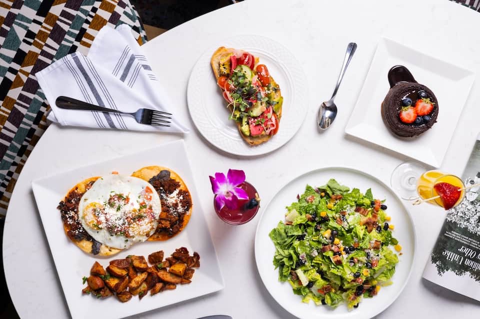 Fine dining spread featuring avocado toast, salad, fruit dessert, and citrus beverages on white plates
