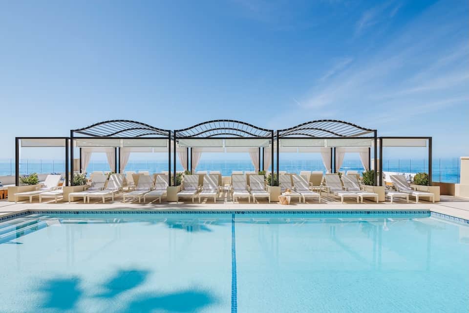Indoor pool with pale loungers under architectural canopies overlooking ocean horizon