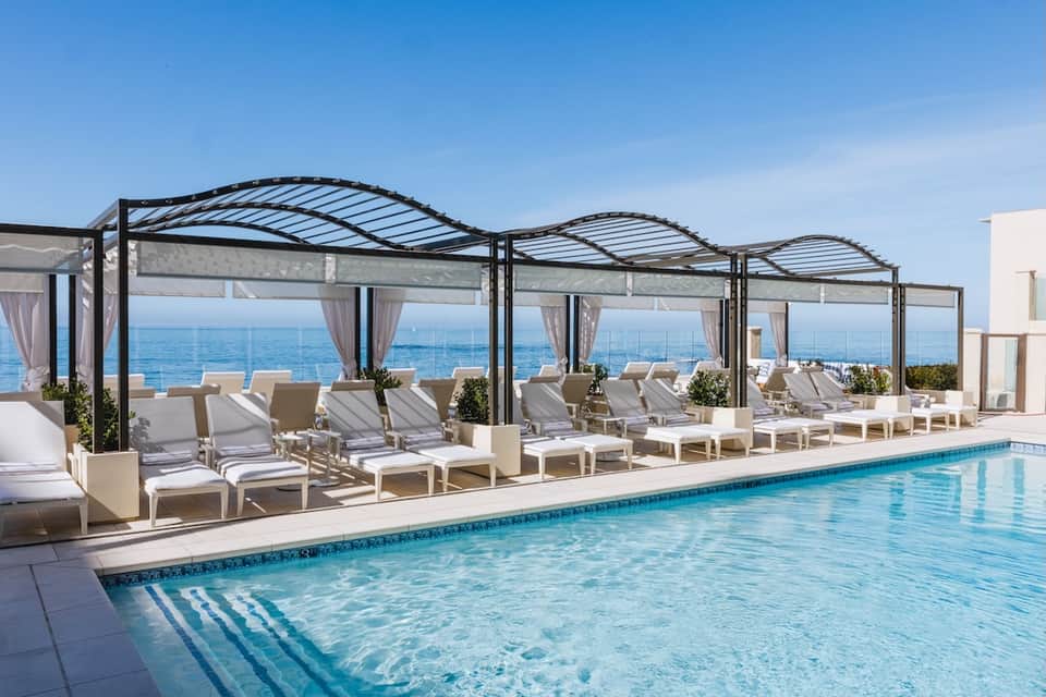 Luxury rooftop pool with ocean horizon, white cabanas, pergolas with shade, and beige lounge seating