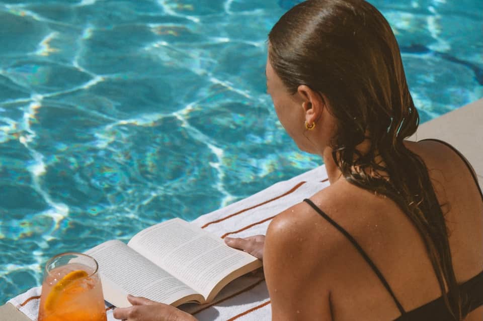 Woman reading poolside while holding a cocktail, overlooking crystal clear turquoise swimming pool water