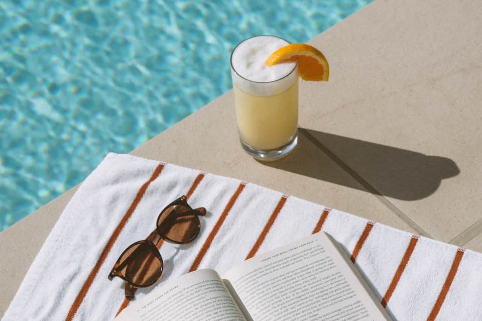 Poolside scene with cold beverage, sunglasses, and open book on striped towel by turquoise water