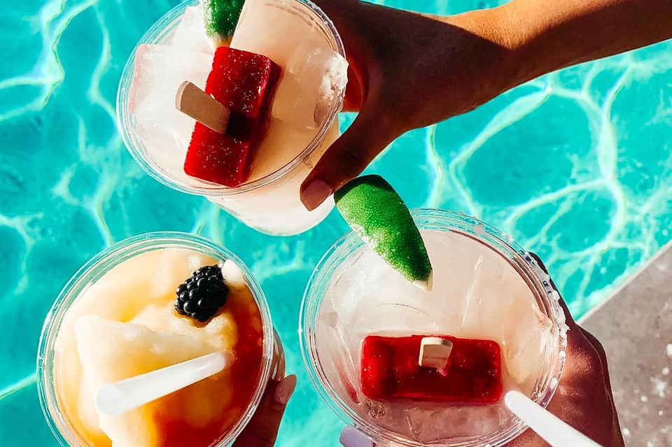 Three tropical cocktails with ice, fruit garnishes, and straws held over turquoise pool water