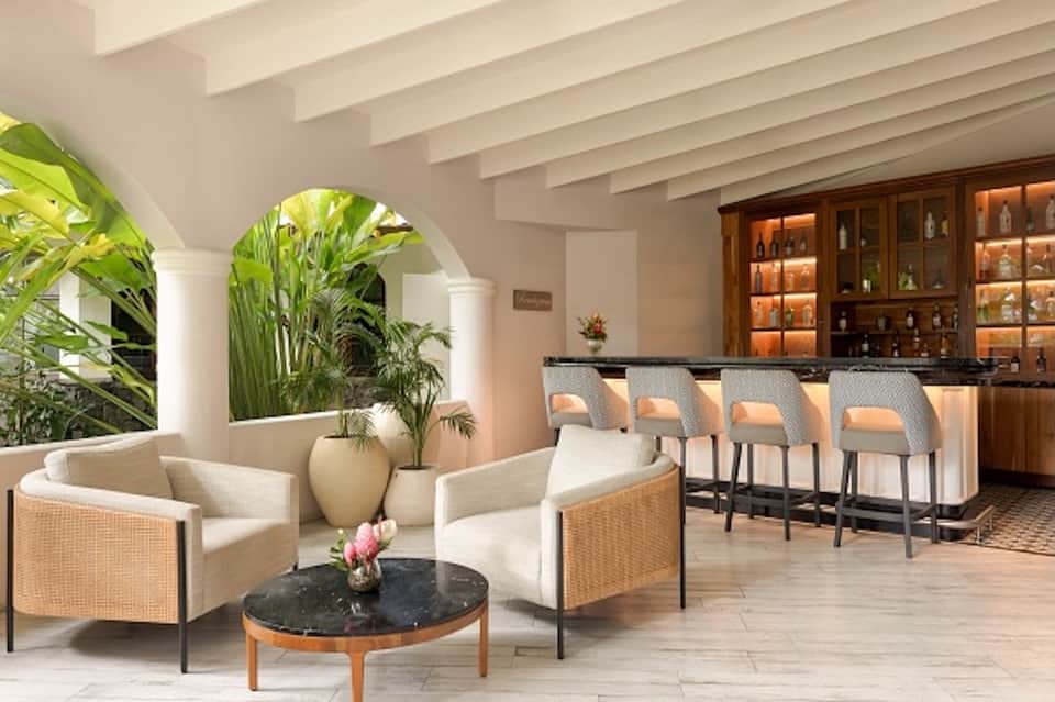 Elegant lobby lounge with wicker chairs, arched columns, tropical plants, and illuminated bar in background