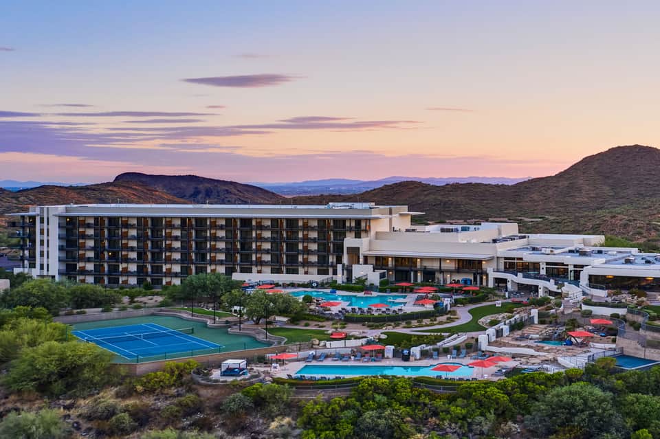 Desert resort at dusk with modern architecture, multiple pools, tennis court, and mountain views