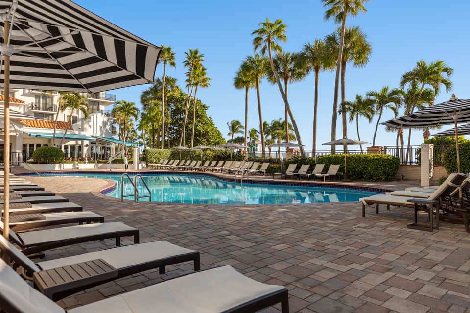 Expansive resort pool with lounge chairs, striped umbrellas, palm trees, and ocean view in background